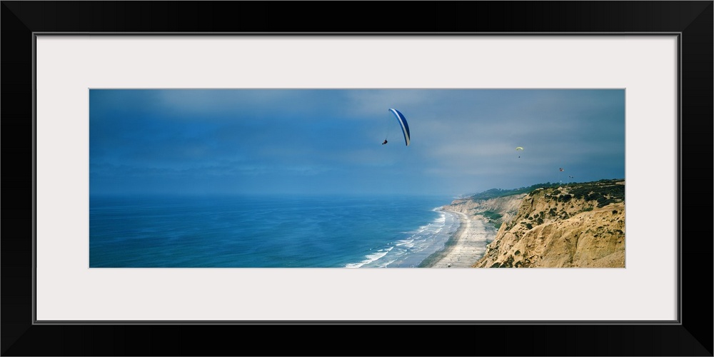 Horizontal photograph on a giant canvas of several paragliders floating over bright blue waters along the coast of La Joll...