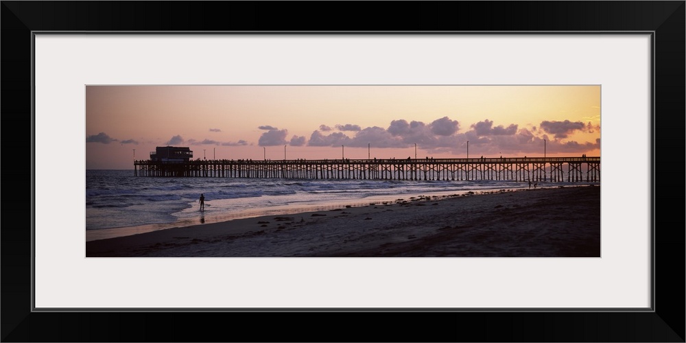 A beach board walk extends out into the ocean in this landscape photograph of the sun setting on a sandy beach.