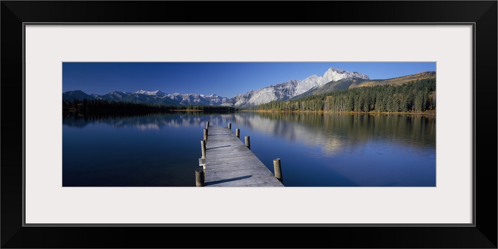 Panoramic photograph of tilted wooden dock stretching into the water.  There is a forest line and range of snow covered mo...