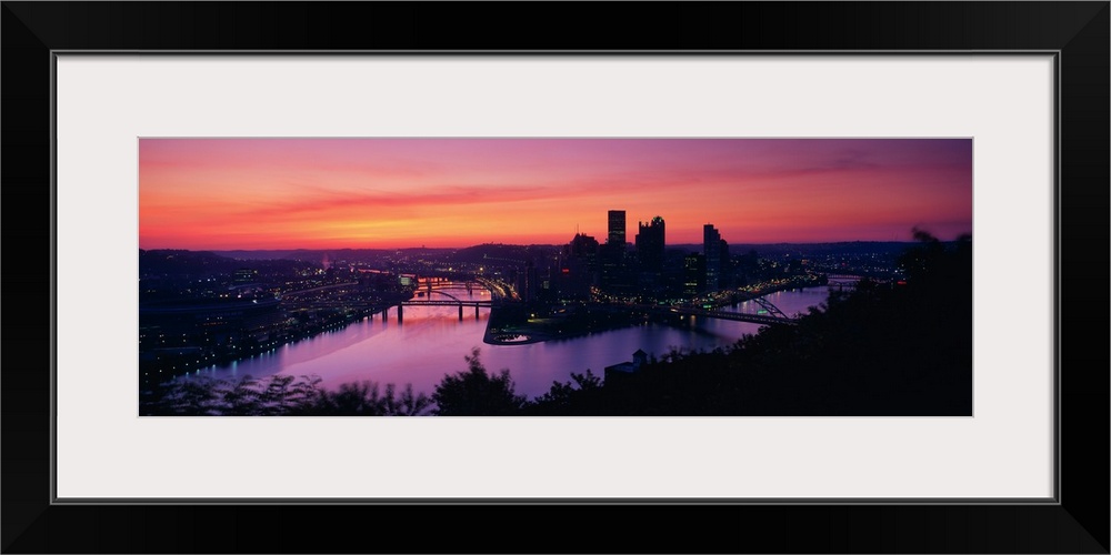 Oversized, landscape photograph of the distant skyline of Pittsburgh, Pennsylvania lit up, beneath a vibrant sunset.