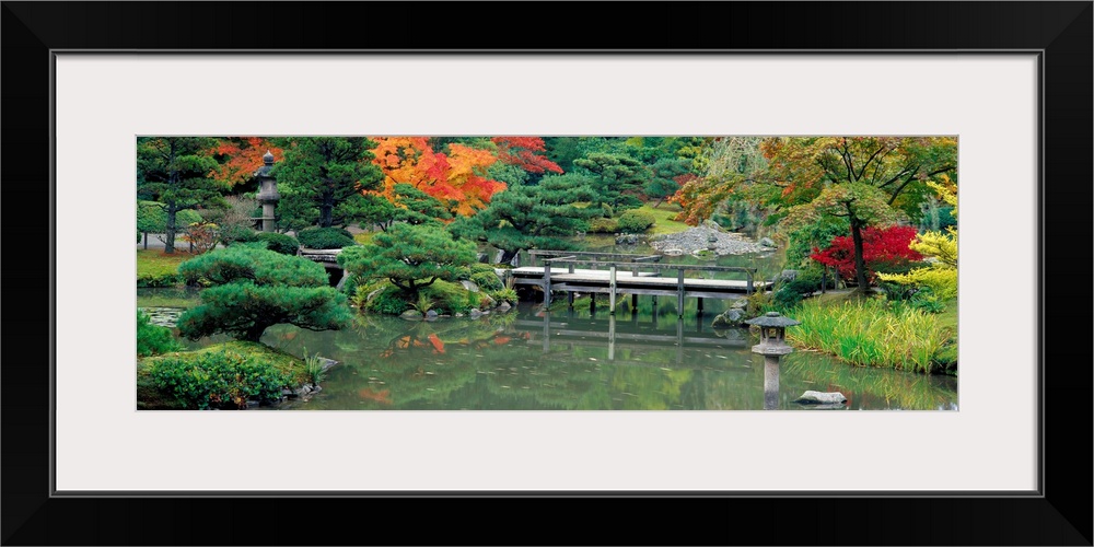 A small foot bridge in a Japanese garden is photographed in a wide angle view with lush green foliage and autumn colored t...