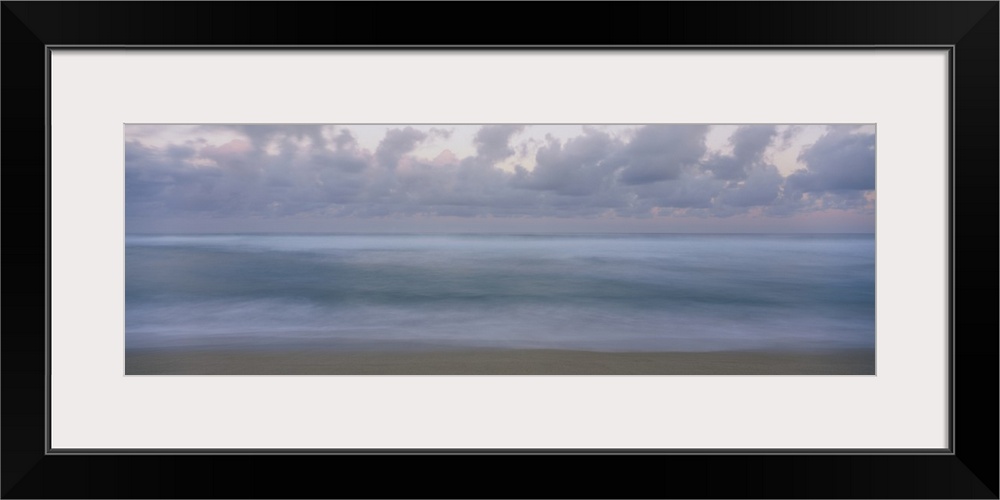 This is a panoramic photograph of a waves gently washing on the sandy shore with a cloudy sky overhead in this seascape.