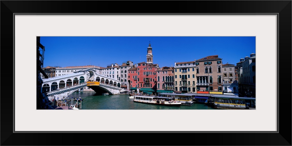 This panoramic view shows the Rialto bridge over the grand canal in Venice with buildings lining the water behind the bridge.