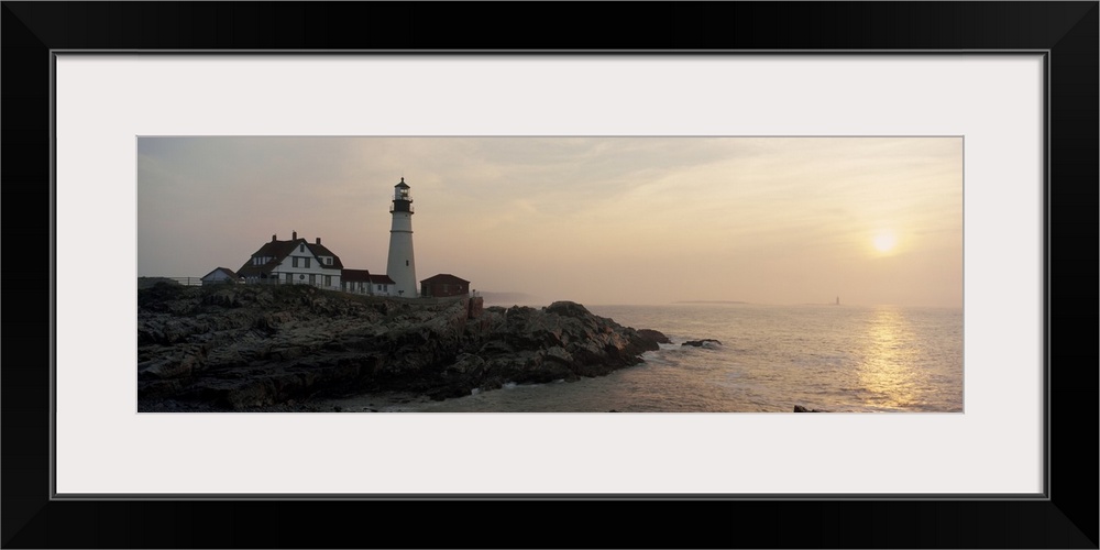 Wide angle photograph of a lighthouse sitting on a cliff that reaches far out into the ocean. The sun has begun to set ove...