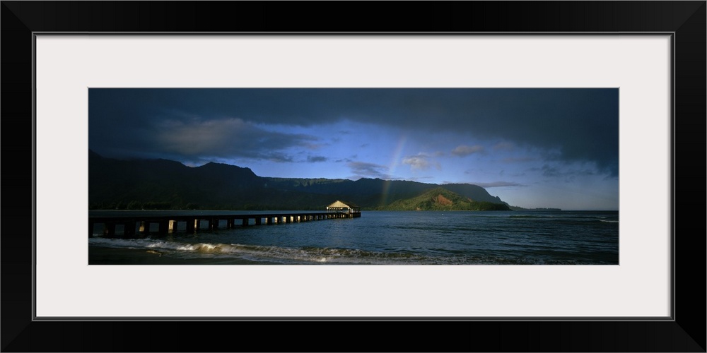 Picture taken of a long pier that reaches far out into the ocean with a faint rainbow shown near land that is in the dista...