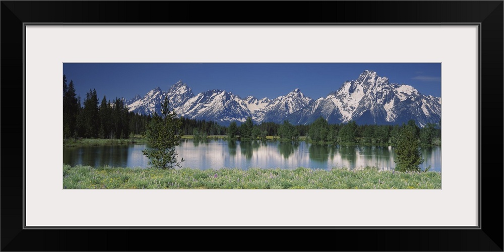 Long canvas of snow capped mountains in the back of a forest reflected onto the waterfront.