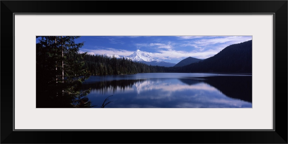 A still lake photographed in wide angle view reflects a mountain that is seen just behind a thick pine forest. The sky fil...