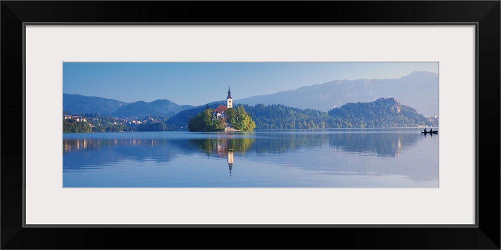 Giant, horizontal photograph of a mountain landscape and buildings reflecting in the waters of Lake Bled, in Slovenia.