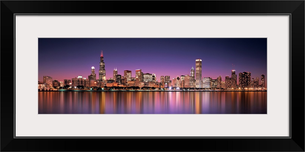 Panoramic photograph of the Chicago, Illinois skyline filled with skyscrapers reflecting onto Lake Michigan at night.
