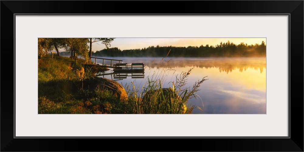 A small dock sits in the water that is lined by tall brush. The sun reflects in the water that has a layer of steam coming...