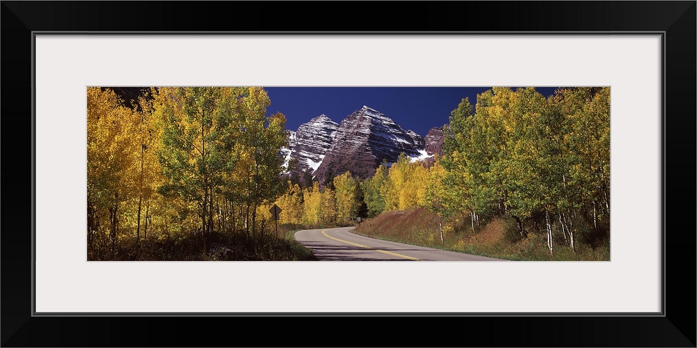 Large, horizontal photograph of a road winding through a fall colored forest, the Maroon Bells in the background beneath a...