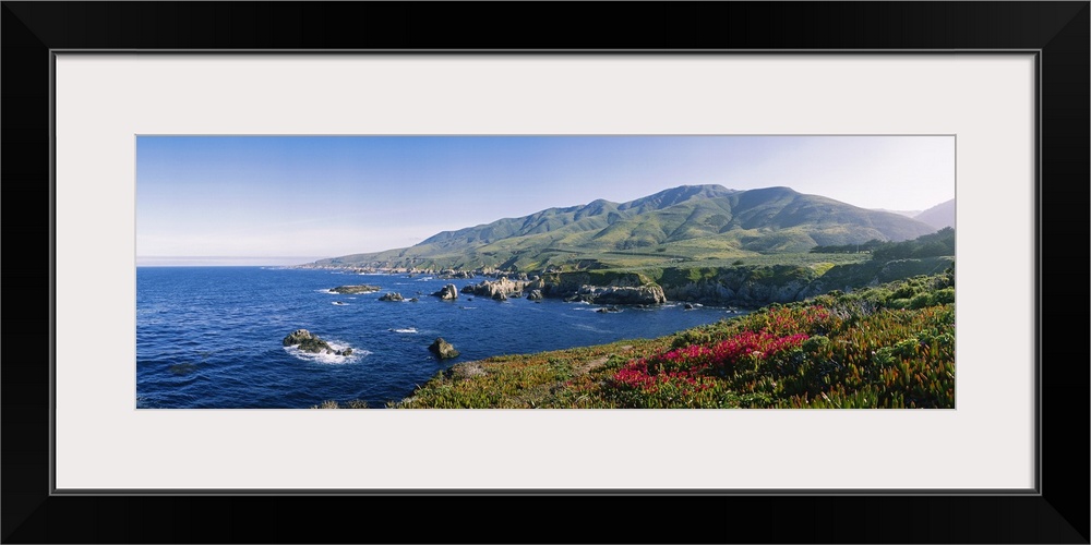 Horizontal, oversized photograph of blue waters with protruding rocks, mountains in the distance, in Carmel, California.