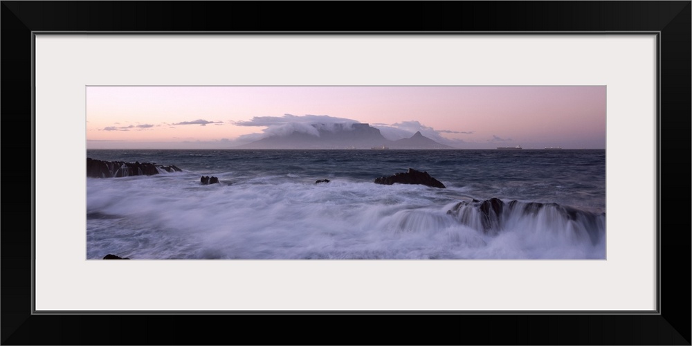 Waves from the rising tide crash over the rocky coastline at twilight. In the distance clouds form over mountain peaks.