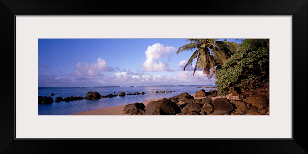 Panoramic photograph displays a sandy beach filled with large rocks and palm trees on a sunny day.  In that distance the t...