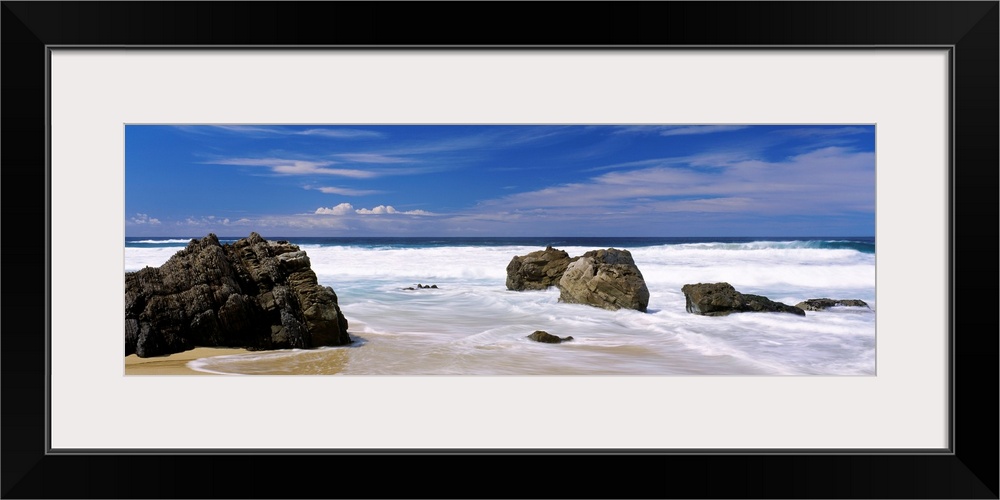 Panoramic photograph of large boulders sitting on a beach in Big Sur, California (CA) on a sunny day as waves trickle onto...