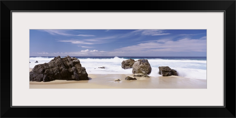 Oversized, horizontal photograph of the big waves of the Pacific Ocean rushing onto a shore with large rocks, beneath a li...