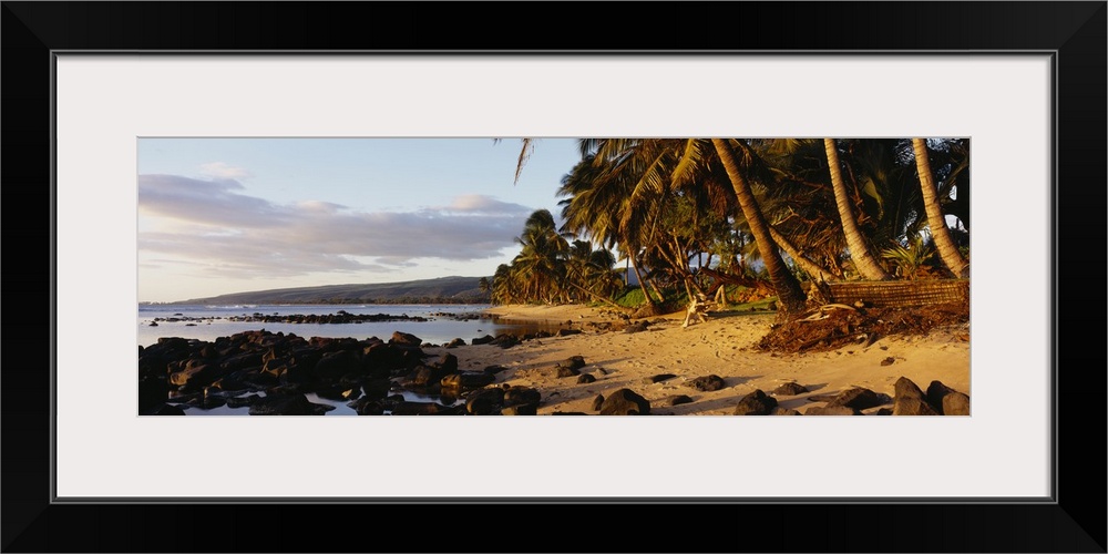 This decorative wall art is a panoramic photograph of sunset on a tropical beach covered with volcanic boulders and lined ...