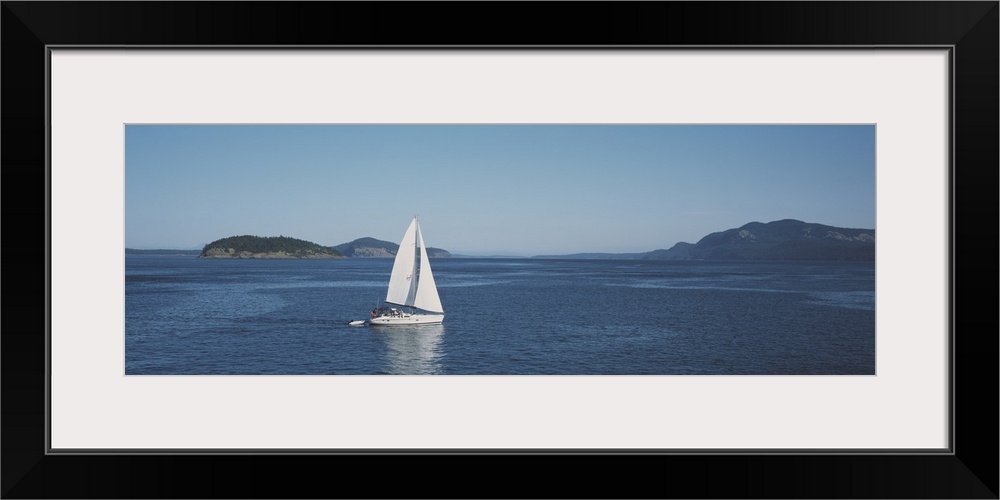 Horizontal and narrow photo on canvas of a sailboat sailing in the ocean with islands covered in forests in the distance.