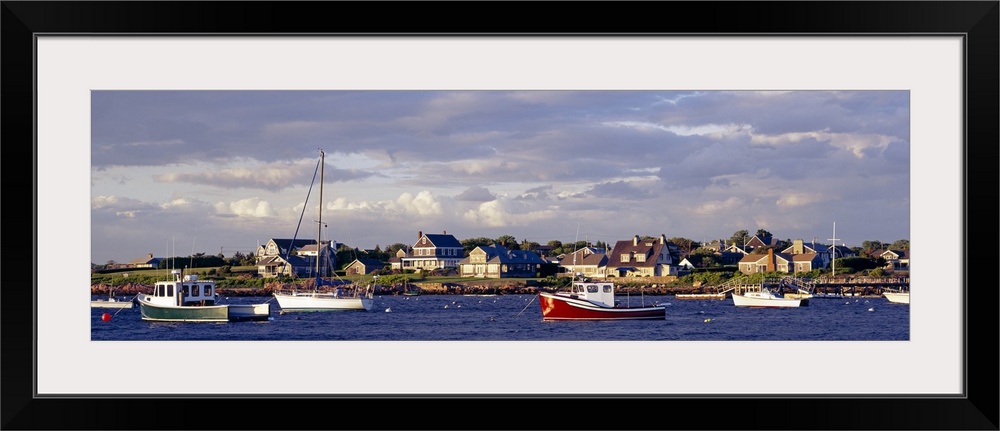 Large landscape photograph of many boast in Sakonnet Harbor, houses line the landscape, beneath a mostly cloudy sky in Rho...