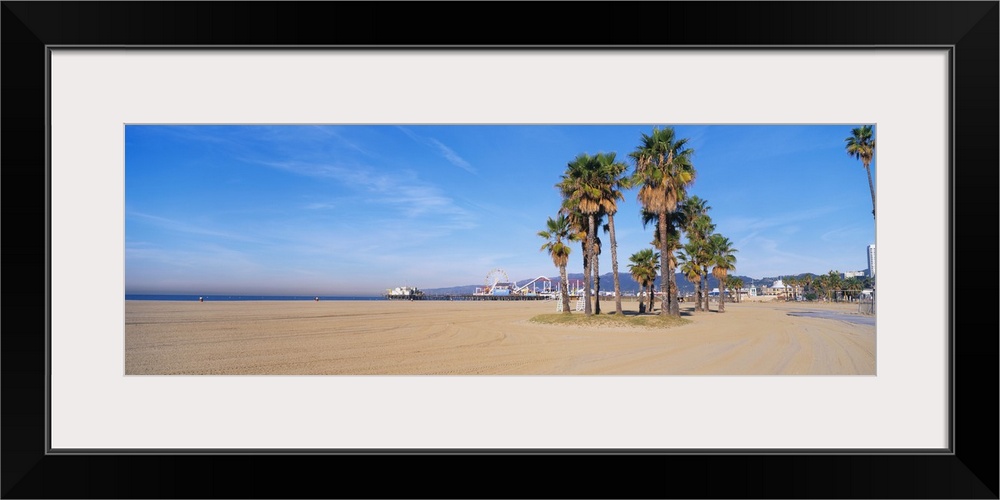 Wide angle photograph taken on a sunny day of Santa Monica beach with a patch of palm trees toward the back near a foot pa...