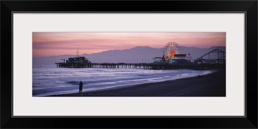 A panoramic shot taken of the Santa Monica pier during sun down with a couple standing on the beach at the waters edge.