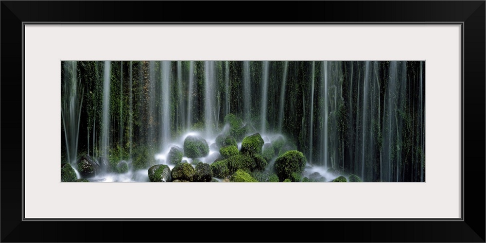 Panoramic photograph displays a gathering of large rocks covered in moss as the waterfall above crashes into and splashes ...