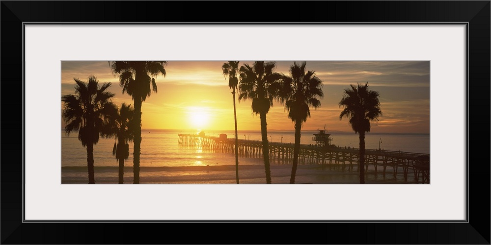 Panoramic photograph of dock stretching into ocean at sunset with palm tree silhouettes in the foreground.