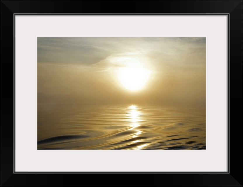 Large landscape photograph of a rising sun covered by a thin haze in the sky, above the rippling, smooth waters of Waterfo...