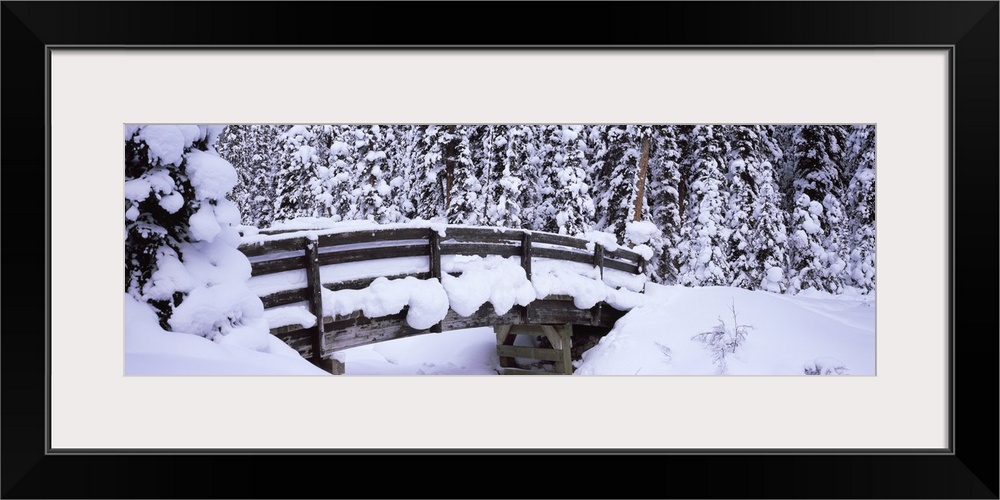 Long horizontal photo on canvas of a heavily snow covered bridge in the middle of a forest.