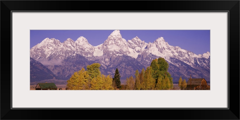 Panoramic photograph taken of an immense mountain range that has the tops covered with snow. Some trees and houses line th...