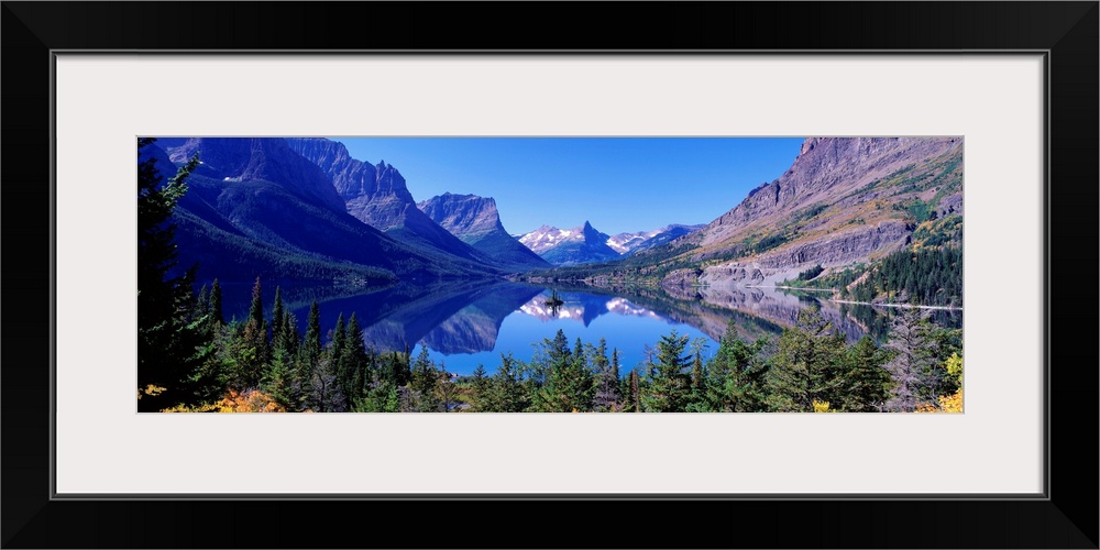 A photographic print of rugged mountains with snow in the distance reflected in a crystal clear lake with foliage in the f...