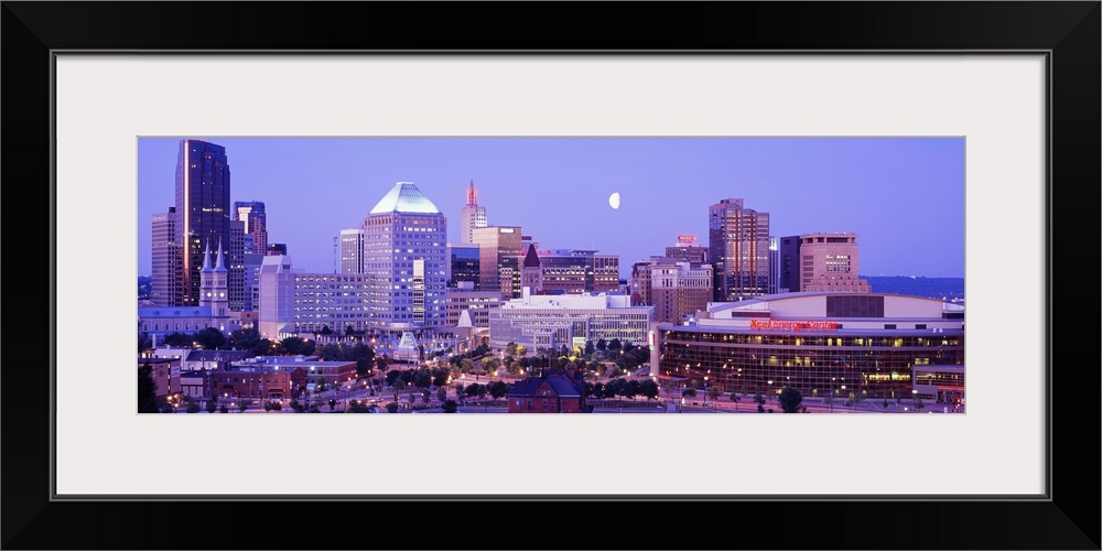 Panoramic photograph of skyline lit up at dusk with moon in the sky.