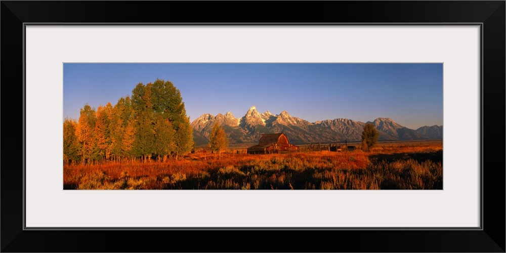 Wide angle photograph of sunrise over a large barn in vast field of tall grasses and trees, mountains on the horizon, in G...