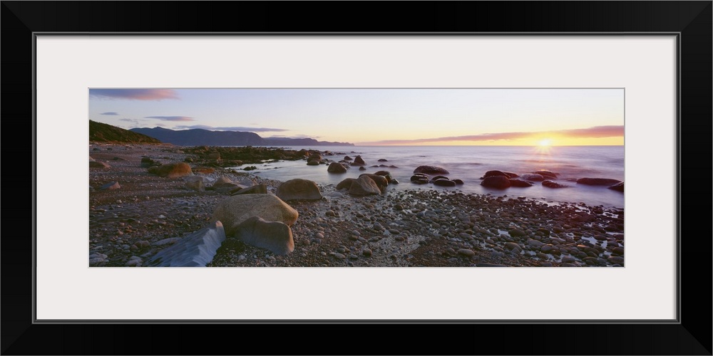 Panoramic photograph of rock and pebble filled shoreline with sun setting in the distance.