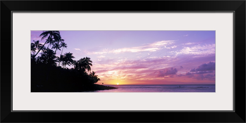 Panoramic photograph displays the sun in the far distance as it begins to set over an island filled with palm trees in the...