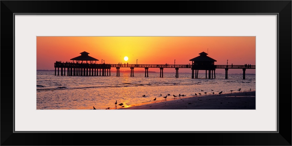 Pier and board walk with shore birds gathered in the foreground as the sun sinks in the sky.