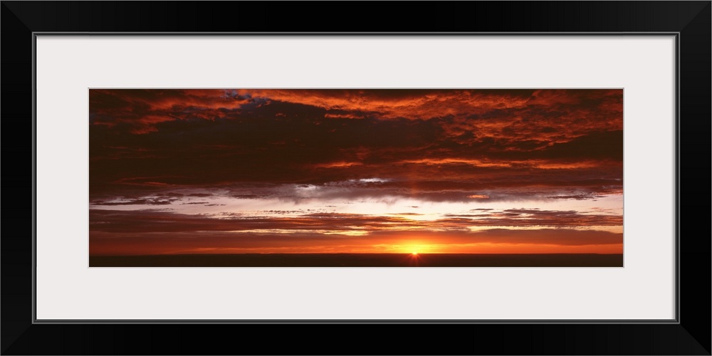 Panoramic photograph of a sunset just hitting the horizon and lighting up a cloud filled sky with warm tones.