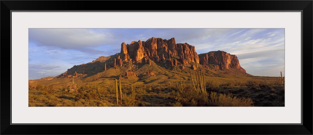 An immense rock formation is photographed from a distance with green land in front of it that is filled with brush and cacti.