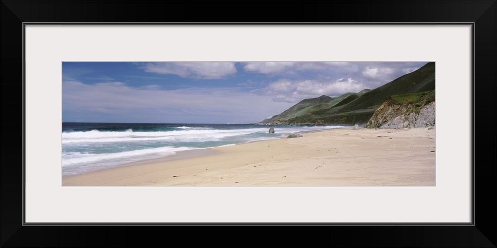 Panoramic photograph of shoreline with mountains.  There are waves rolling in under a cloudy sky.