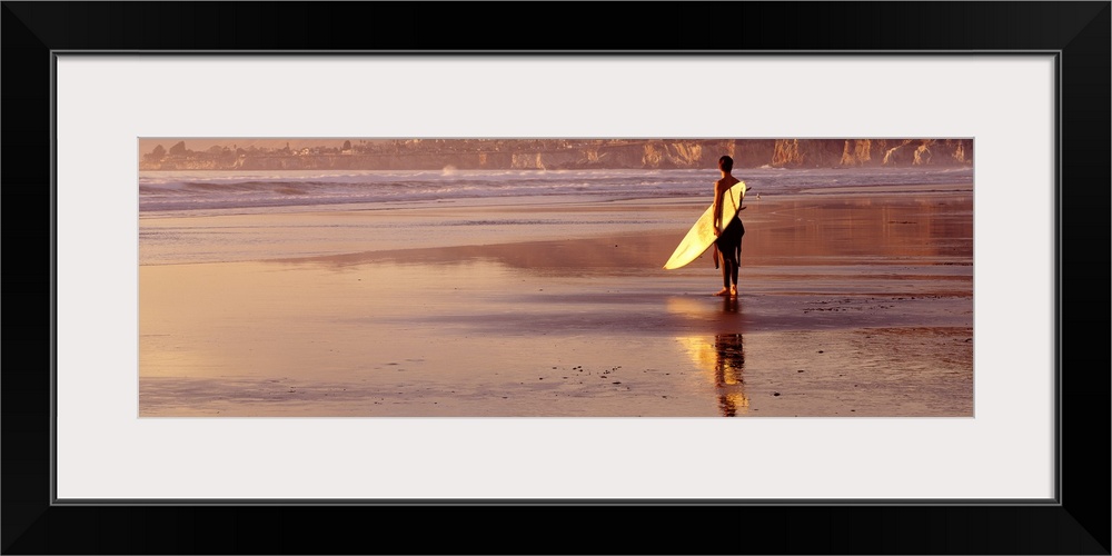 Panoramic photograph of a single surfer walking along the shore with his surfboard at Pismo Beach, California.