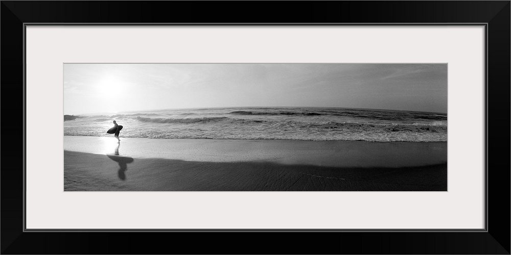 Panoramic photograph of a surfer walking along a sandy beach in San Diego, California.  The tide of the ocean gently crash...