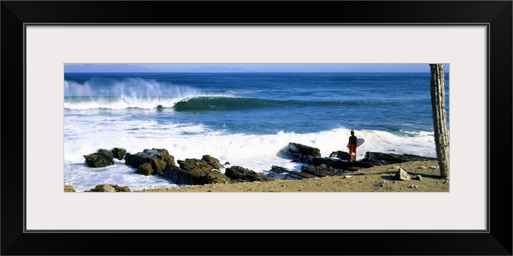 A single figure stands amongst rocks watching waves break on the shore in this panoramic photograph.