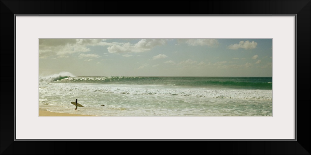 Panoramic image of a surfer standing where the ocean meets the beach shore looking at a big wave crashing.