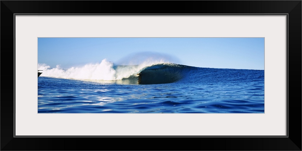 Large, horizontal photograph of a distant surfer riding a large wave, in the blue waters of Tahiti, French Polynesia.