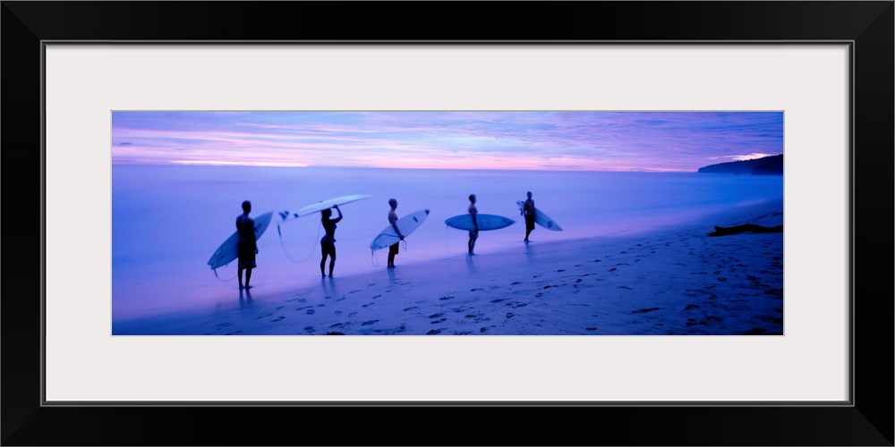 Panoramic photograph shows a group of five surfboarders walking along a sandy shoreline under dim light in Central America.