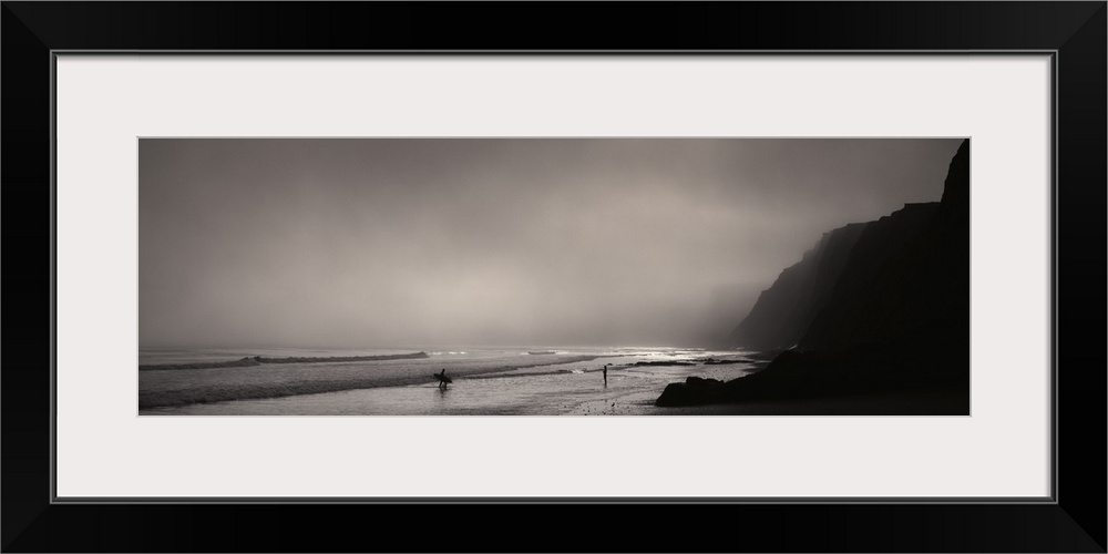 Panoramic photograph on a giant canvas of a dark, foggy sky over Point Reyes National Seashore, where two surfers walk awa...