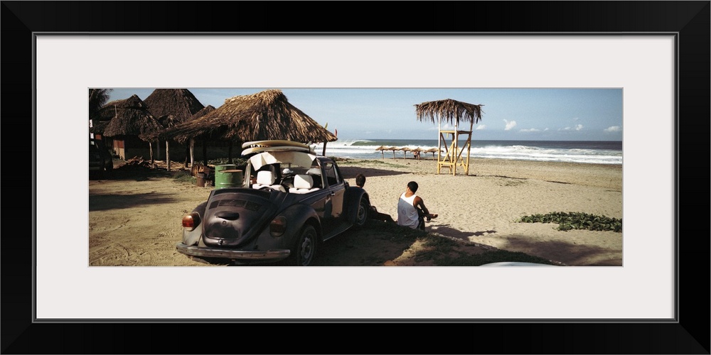 A big panoramic photograph of surfers sitting in front of their car looking out at the ocean. Small beach huts are just to...