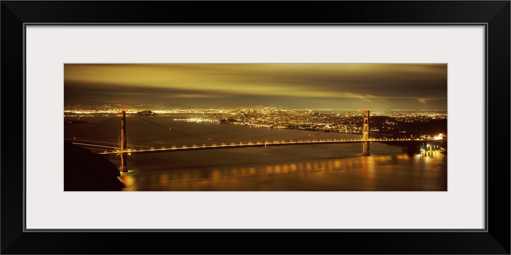 Giant, wide angle photograph of the Golden Gate Bridge lit up at night, the city lights of San Francisco in the background.