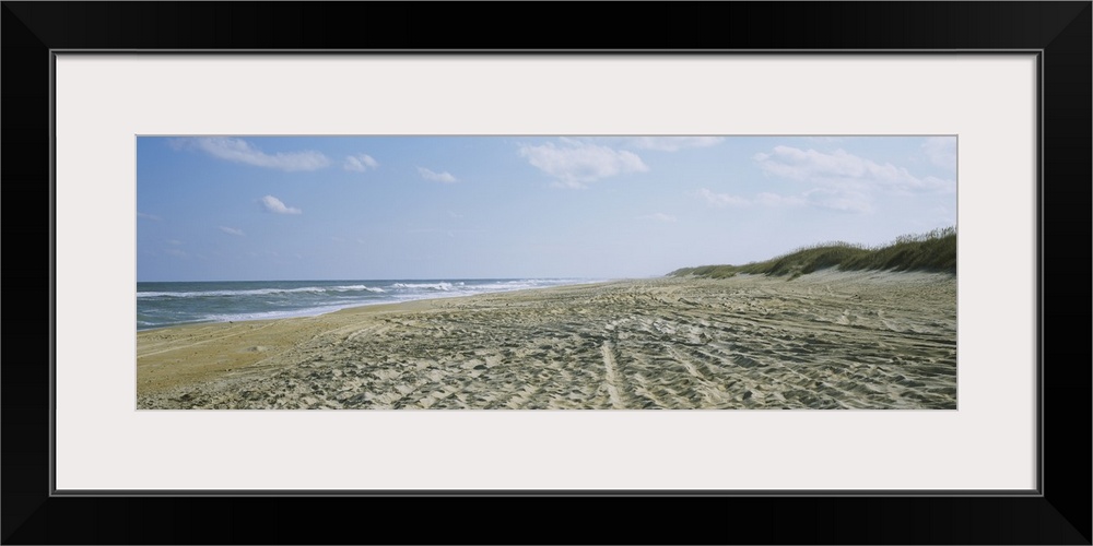A panoramic photograph of a sandy beach indented with footsteps and tracks down to the ocean water.