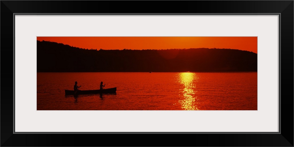 This panoramic piece is a picture taken of two people canoeing on a lake during sunset which has silhouetted the trees in ...